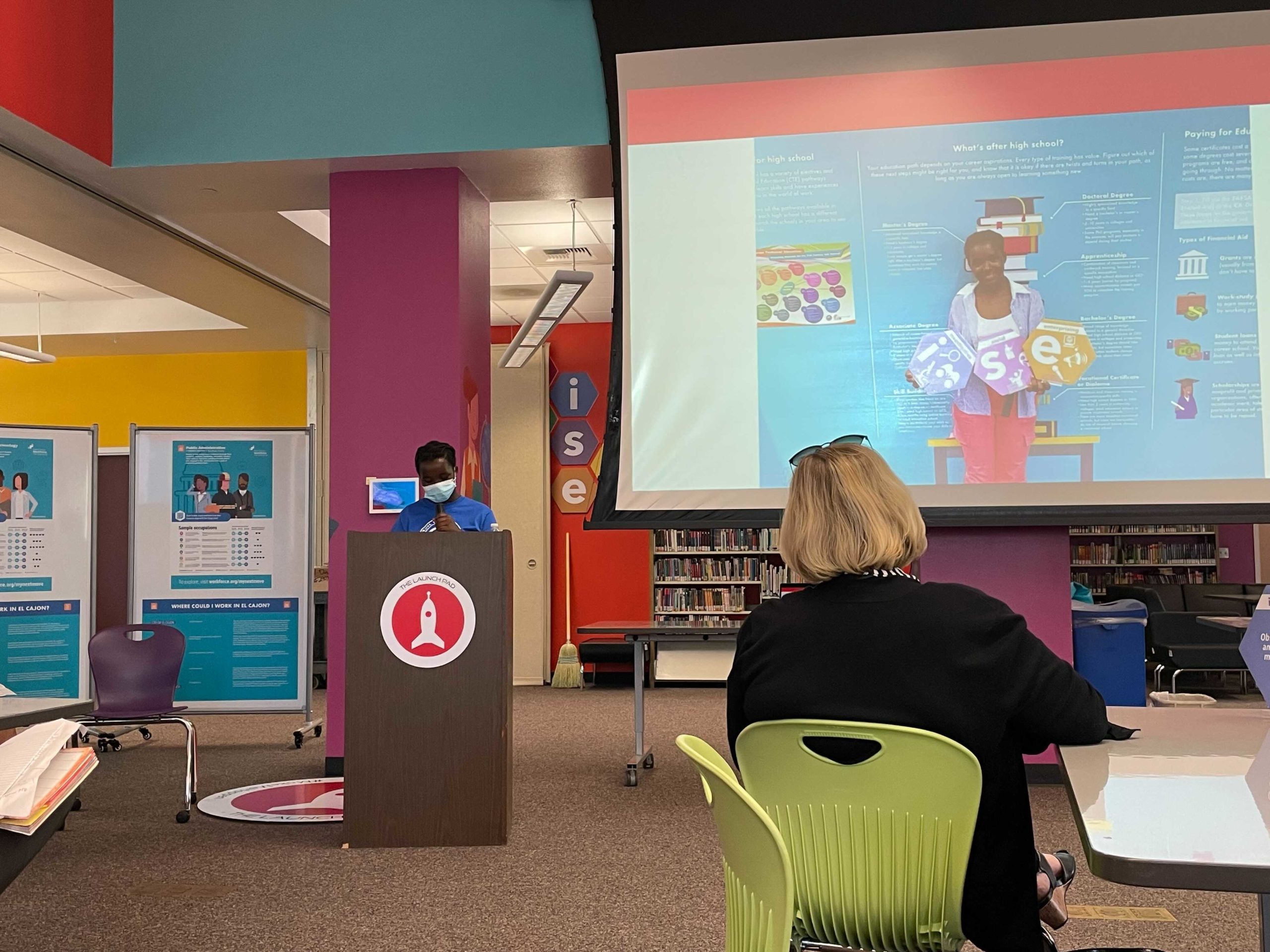 Young black woman stands at a podium wearing a face mask and presents next to projector with photo of her