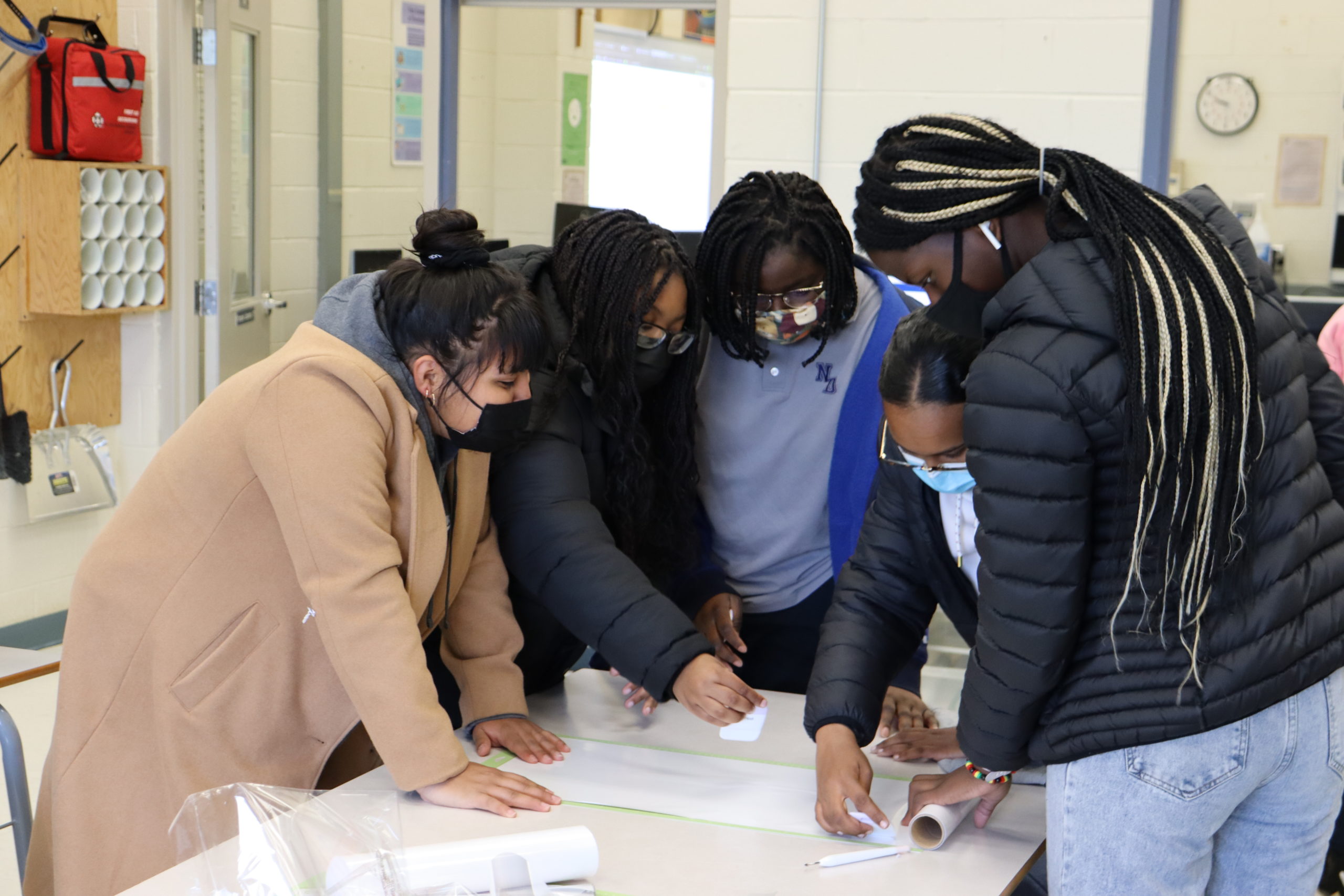 Alt Text: Students standing around a table taking notes on a piece of chart paper. 