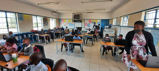Students sitting in desks and a teacher standing at the front of the room on a laptop.