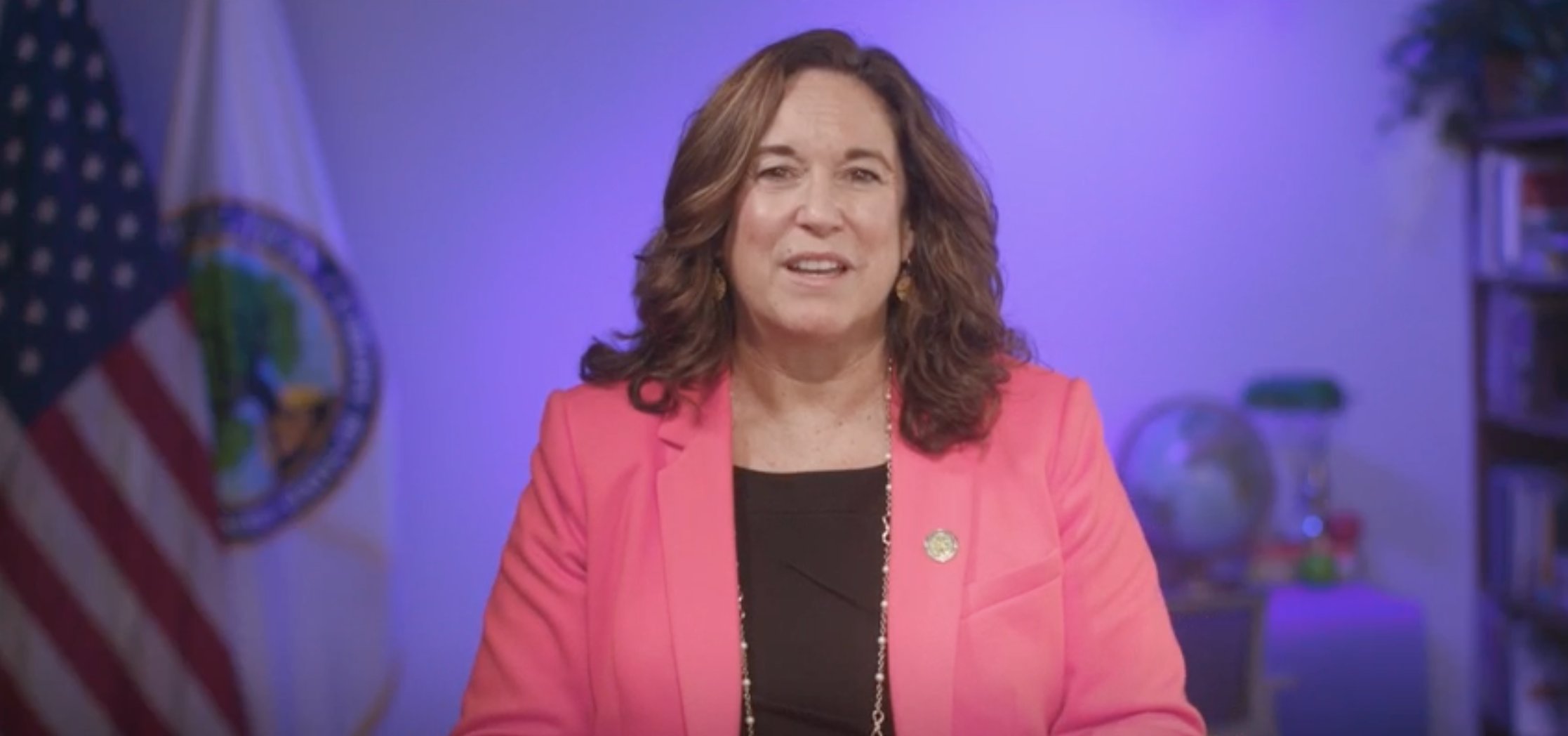 Woman wearing a pink blazer standing in front of a purple background with an American flag and U.S. Department of Education flag behind her