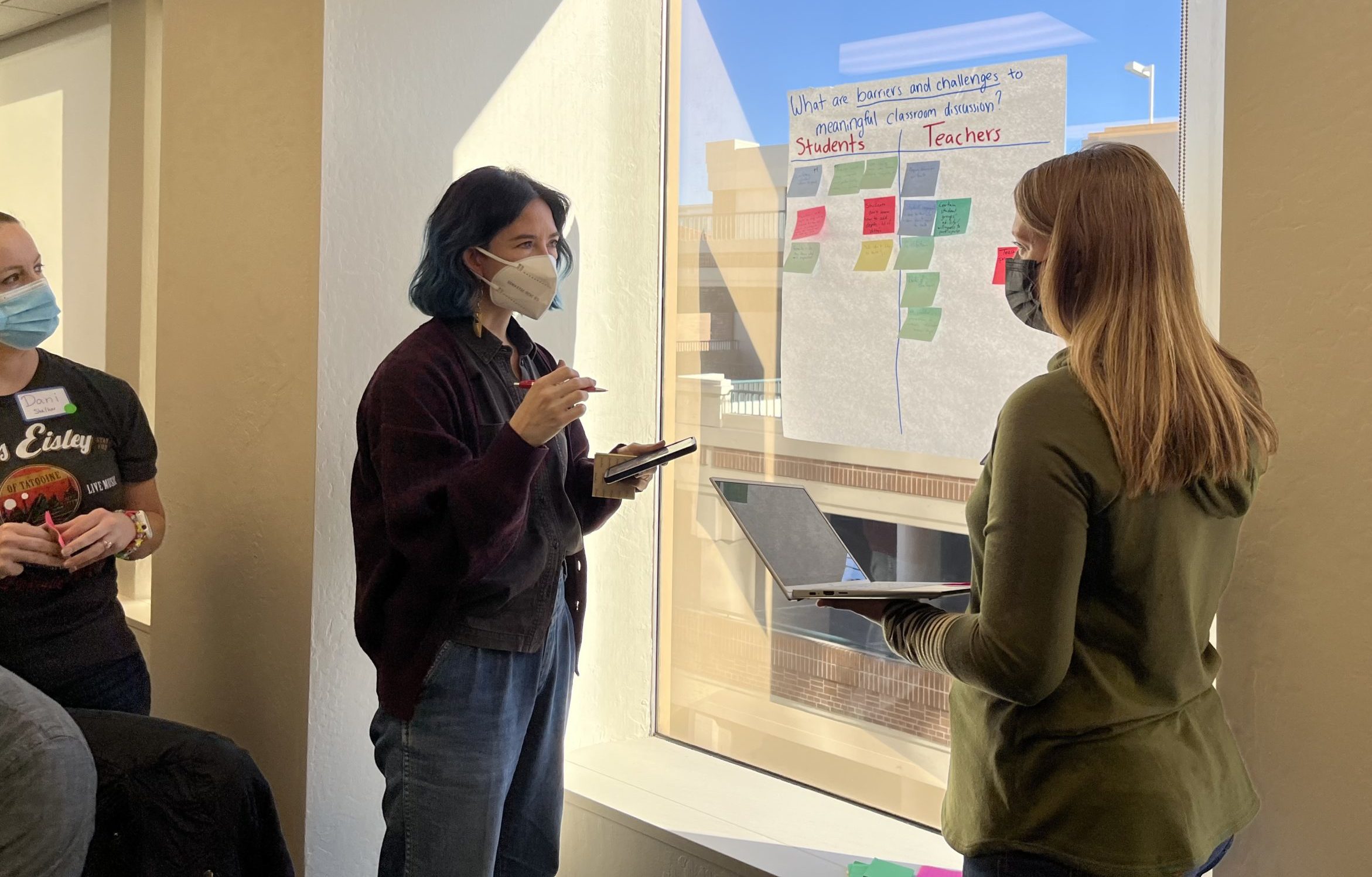 An image of three people talking and drafting notes on post-its, while sticking them to a large poster that is on the wall