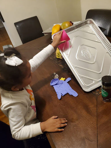 A girl rolls a pink cup down an aluminum foil pan, which is propped up like a slide.
