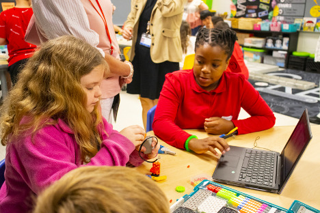 Two female students sit at a table in front of a laptop and robotics kit. The white, female student in the forefront sets up the motor.