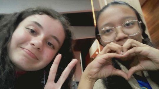 Students Aline M. and Havelline D. take a selfie, making a peace sign and a heart sign with their hands.