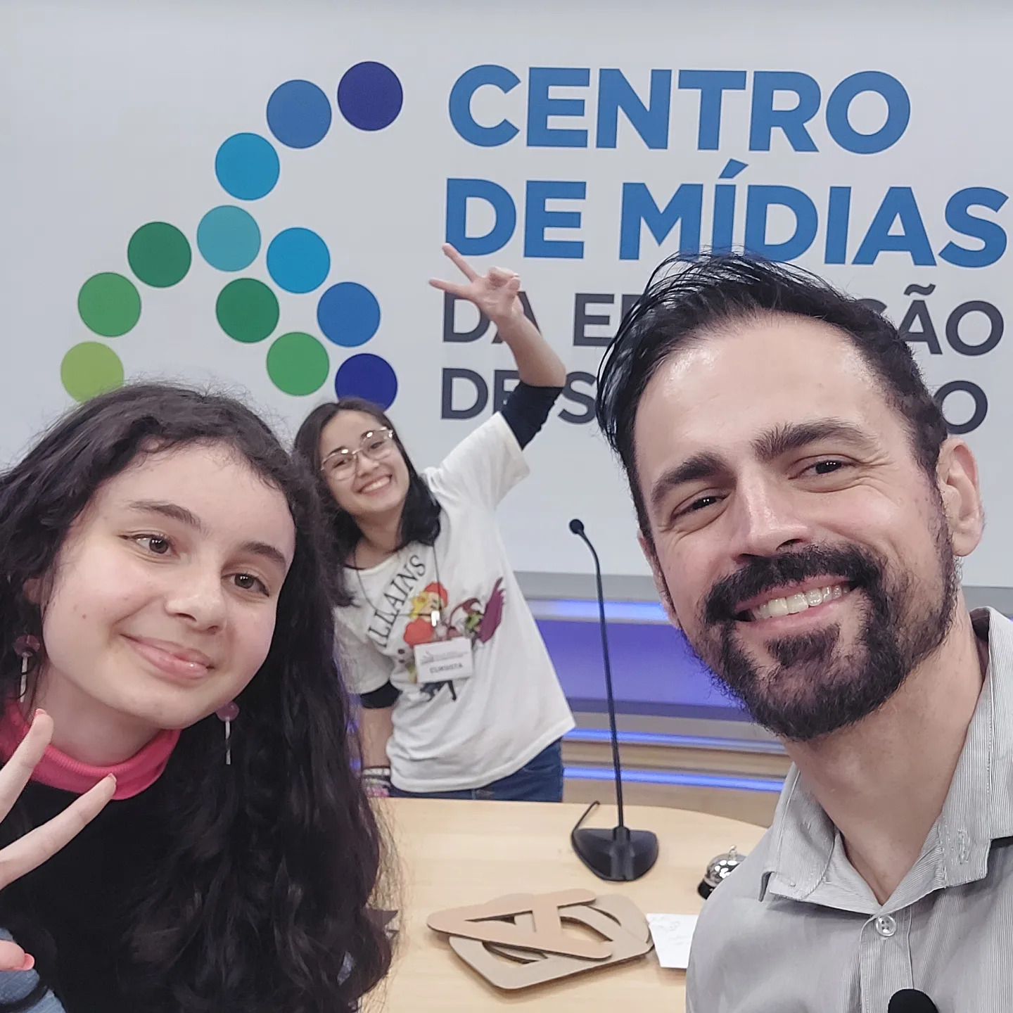 Students Aline M. and Natália C. with teacher Ed Gomes Jr. take a selfie in front of a Centro de Mídias sign