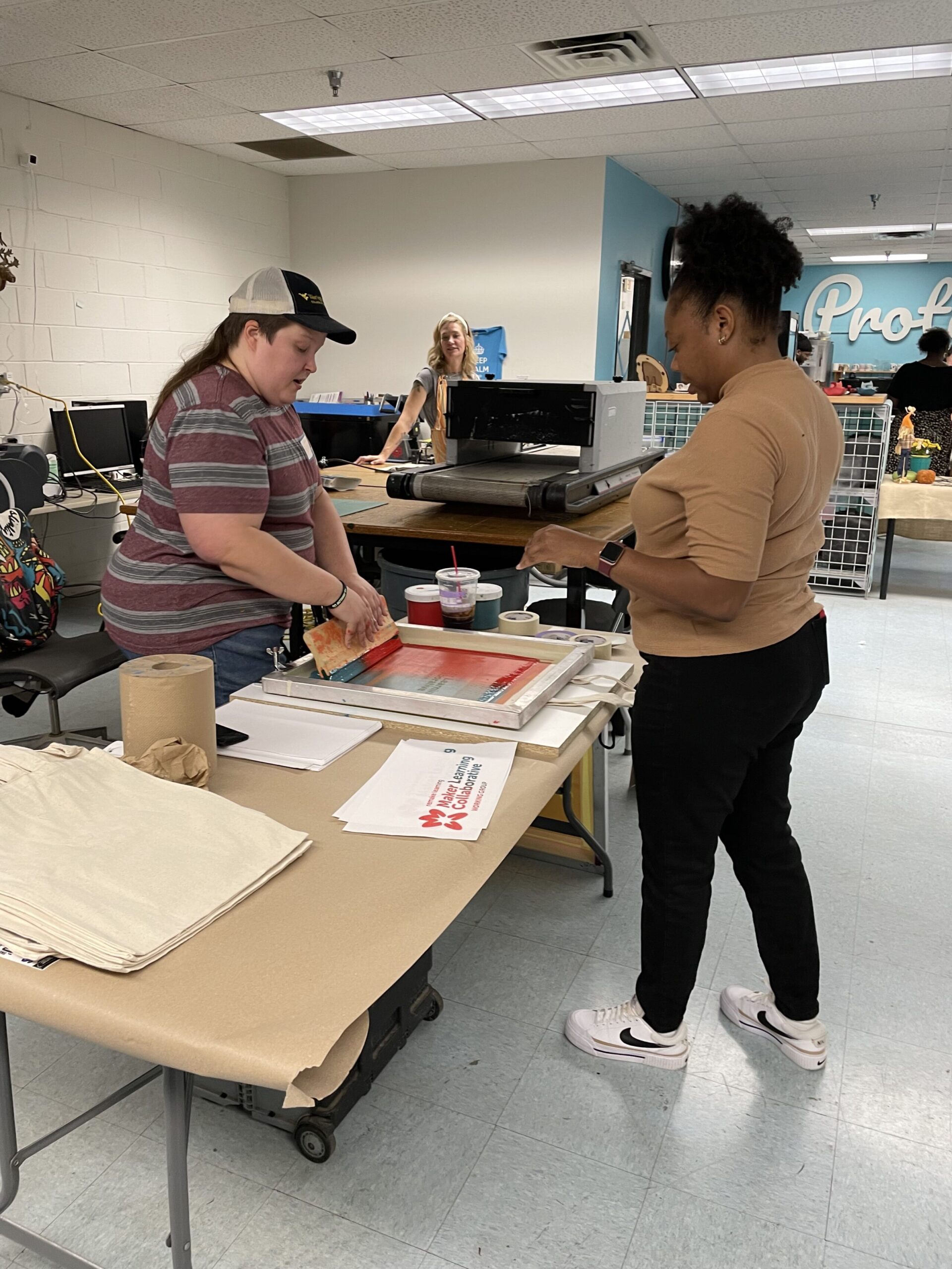 A woman is standing at a table watching the screen printing process of paint being squeegeed through a screen with the Maker Learning Collaborative logo onto a natural canvas tote bag. 