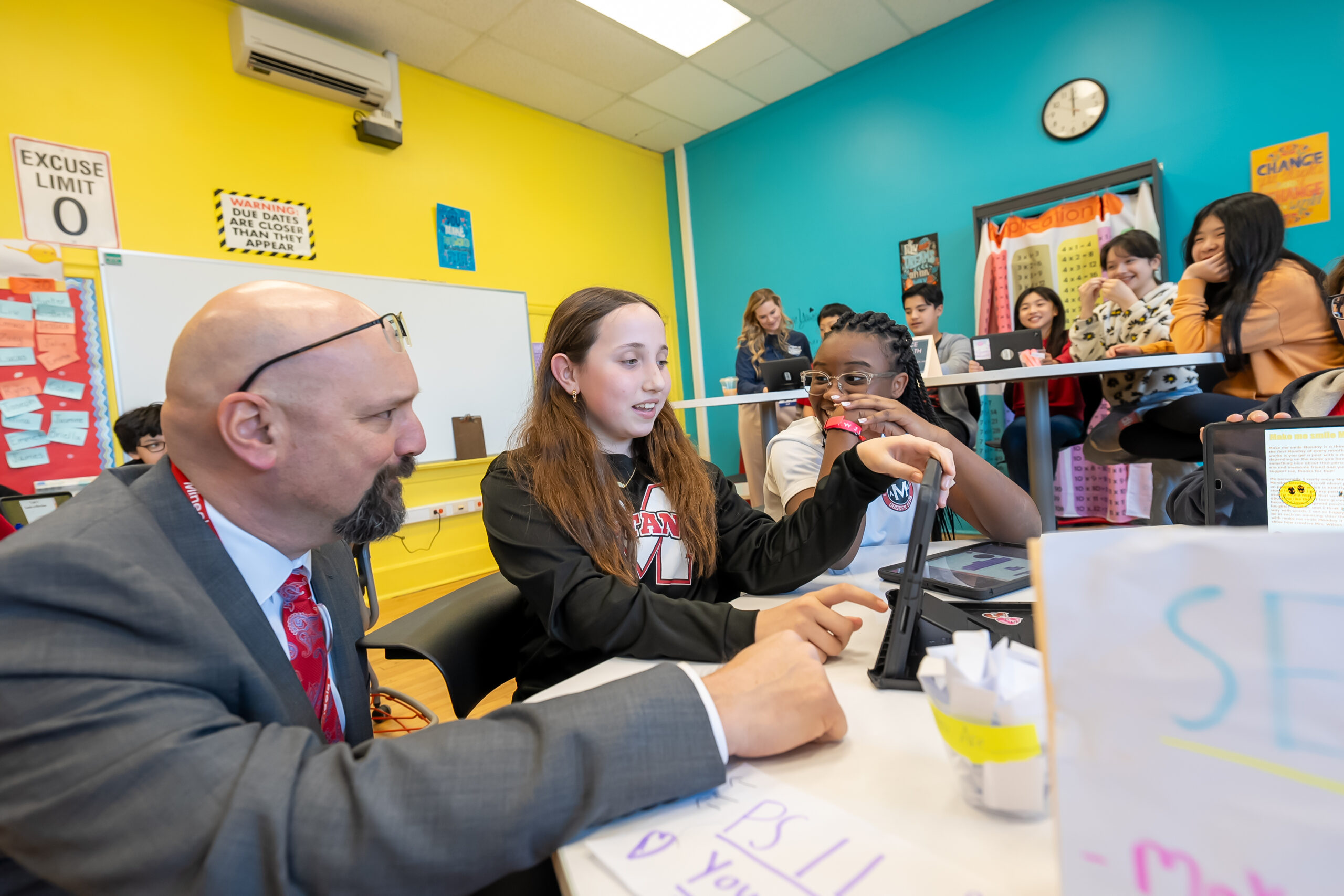 A man kneels beside a table with two young students, looking at a tablet. 