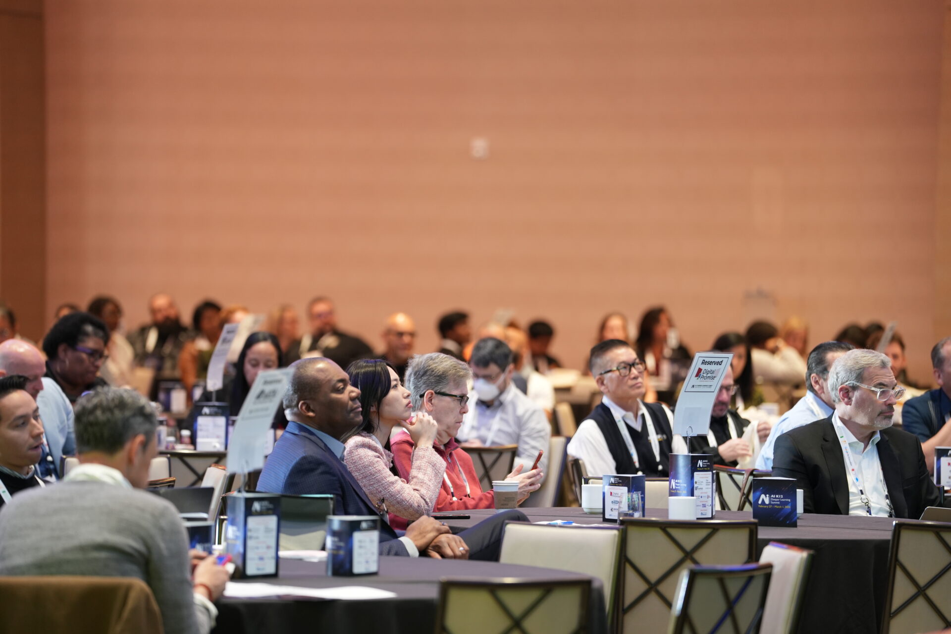 A large group of people sit at tables at a conference, looking to the front of the room. 