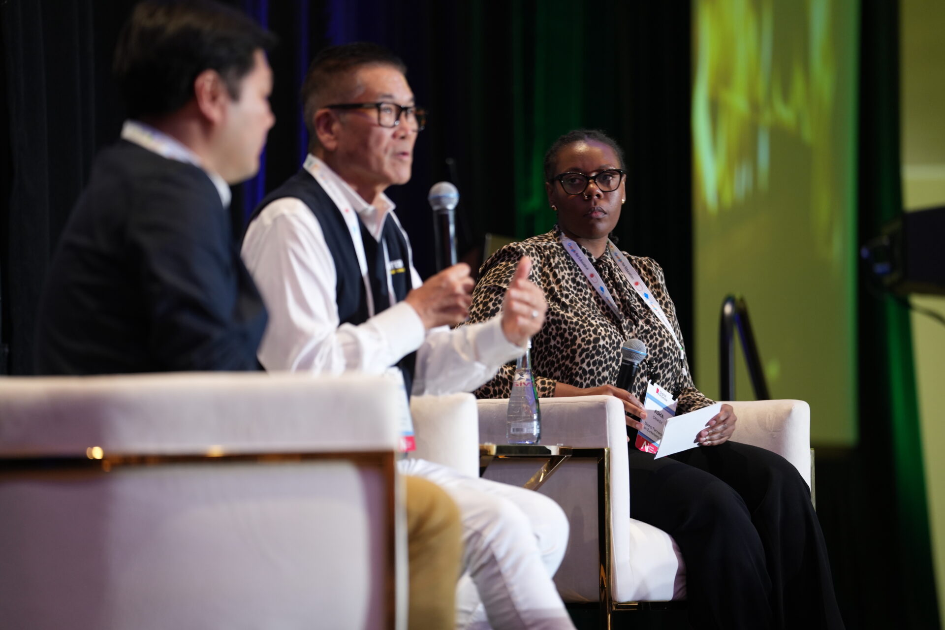 Three speakers sit on chairs on a stage. One man in the middle speaks into a microphone. 