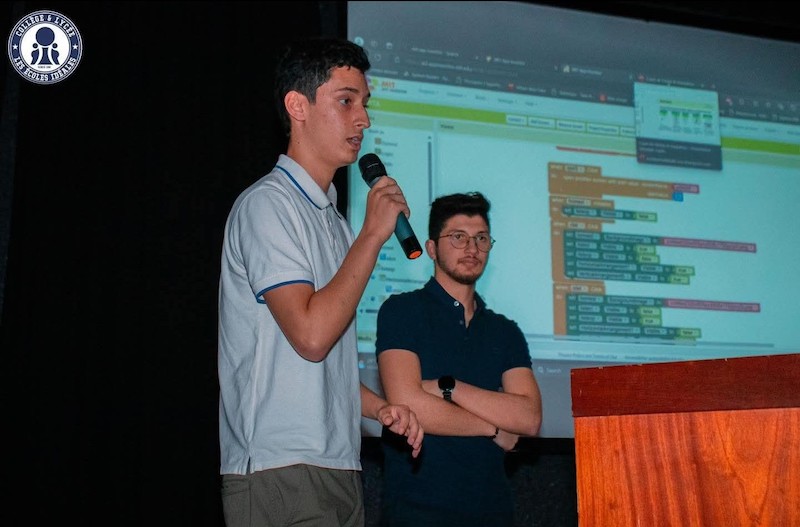 One student speaks into a microphone on stage while another student stands next to the screen
