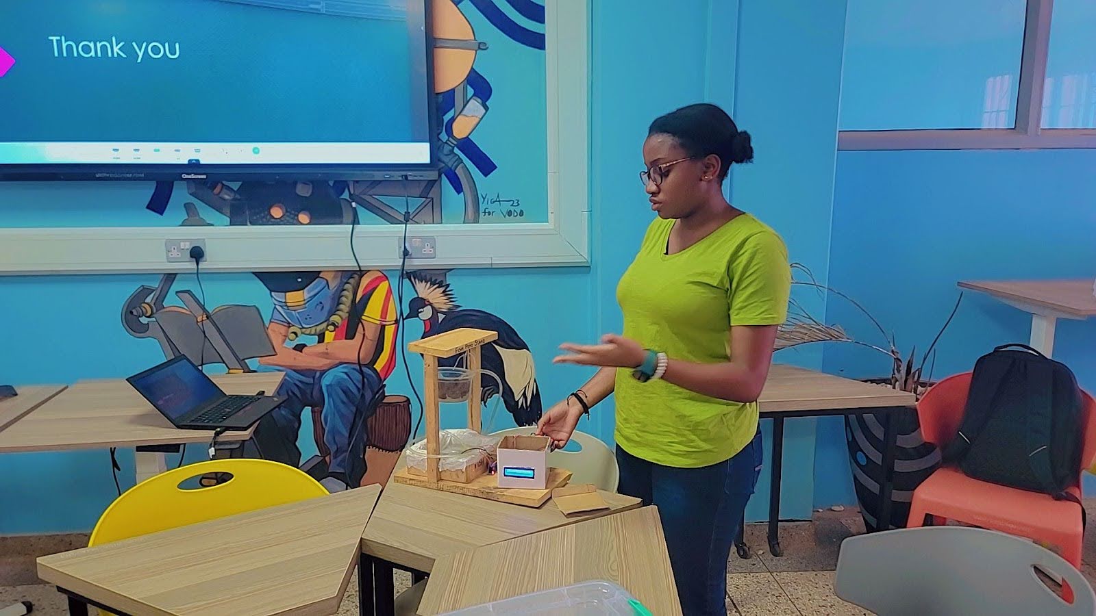 A student presenting a school project model on a table in a classroom with vibrant blue wall murals.