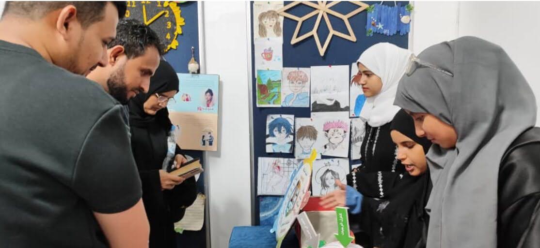 Three students presenting their project to observers at a community event surrounded by student art.